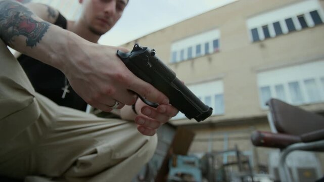 Low angle close up shot of tattooed man crouching outdoors and reloading handgun