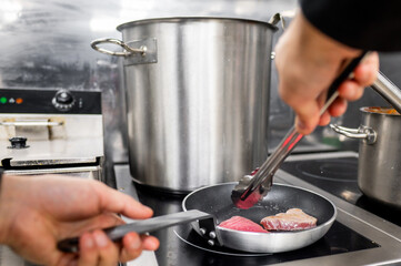 A close-up of a hand using tongs to cook tuna in a frying pan on a stove, with a large pot in the background. The scene captures the essence of home cooking and culinary preparation