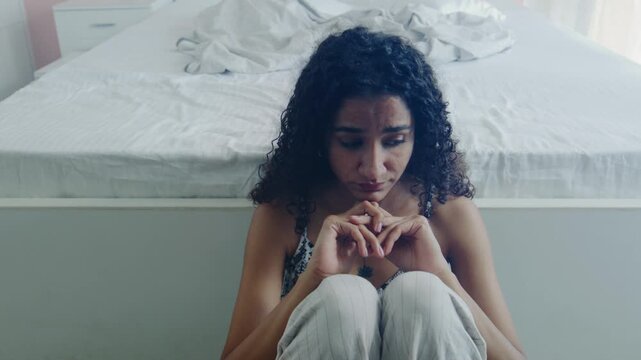 A wide shot of the young curly-haired pensive lonely anxious Indian Asian female or woman sitting on the floor in destress in the bedroom. Concept of heartbreak, depression, mental health medication