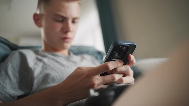 Caucasian teenager chats in messaging app with friends or watches content using mobile phone while sitting on sofa in living room. Teen boy spending weekend at home. Lifestyle concept. Close Up.