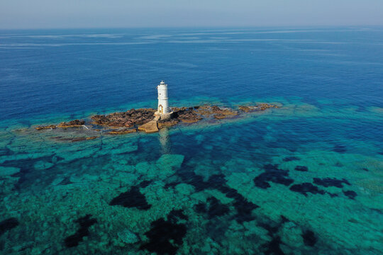 Vista aerea mozzafiato del faro Mangiabarche di Calasetta, Sardegna, Italia. Il faro bianco si erge maestoso su uno scoglio circondato da acque cristalline che sfumano dal turchese allo smeraldo