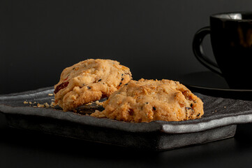 Delicious cookies contain dried fruits or berries like cranberries or raisins, giving them a rustic, homemade look on a ceramic plate against a black background and a teacup.