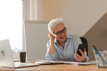 Confident middle aged senior woman euphoric winner with smartphone. Older mature lady looking at...