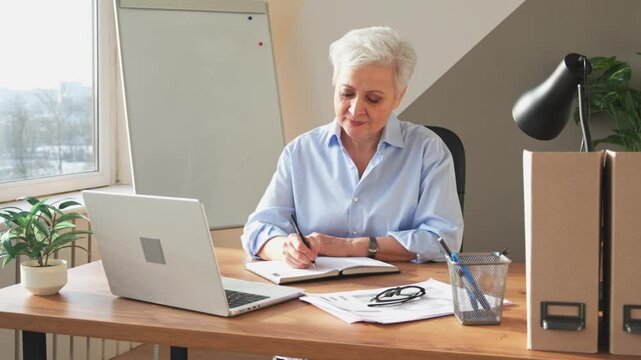 Middle aged senior woman using laptop computer writing notes at workplace. Focused mature old businesswoman work at office making written records doing online work. Mature boss professional worker