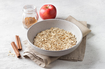 Raw rolled oats in a ceramic bowl with cinnamon sticks and apples on a light gray textured background