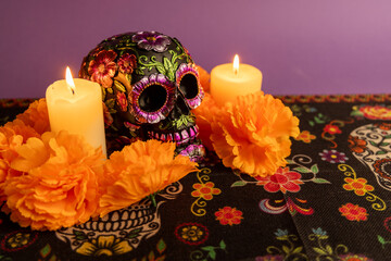 An ornate skull with metallic flowers, flanked by two lit candles. Orange flowers encircle the...