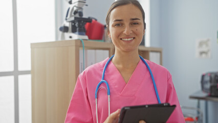 A young woman nurse wearing a pink uniform and a stethoscope smiles while holding a tablet in a...
