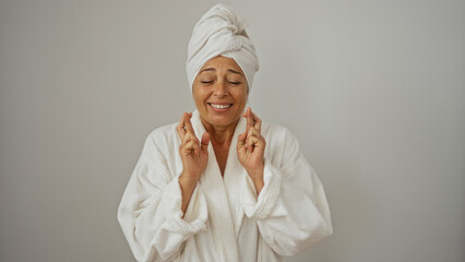 Beautiful elderly hispanic woman with a towel on her head and fingers crossed wearing a bathrobe isolated on a white background