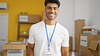 A smiling, bearded hispanic man in a white t-shirt, wearing a 'volunteer' lanyard, stands in a warehouse filled with donation boxes. © Krakenimages.com