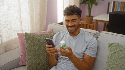 Young man indoors smiling at his phone while holding a pill bottle, seated on a couch in a cozy and...