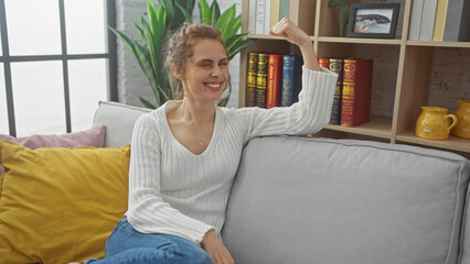A smiling caucasian woman flexing her arm muscles while sitting on a couch indoors, conveying...