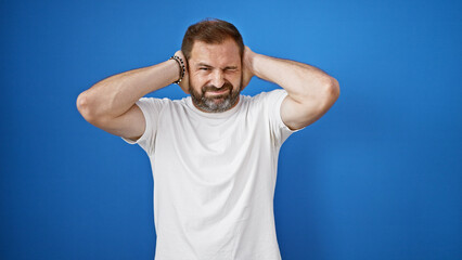 Mature hispanic man with grey hair covering his ears with hands against blue background, portraying frustration or noise.