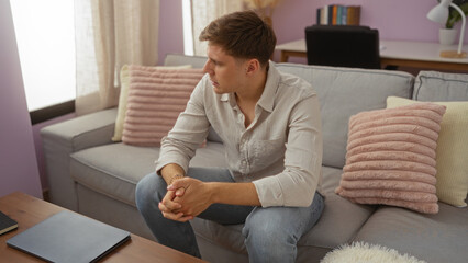 Young man sitting on a gray couch in a cozy living room, looking thoughtful and relaxed in casual attire, surrounded by pillows and pastel-colored decor.