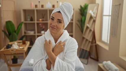 Woman relaxing at a spa in a white bathrobe and towel wrapped around her head, standing in a serene indoor wellness room with plants and wooden shelves in the background