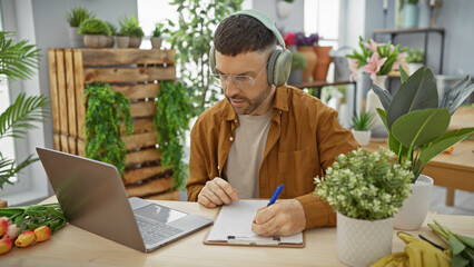 Fototapeta premium A young hispanic man with a beard works attentively on a notepad at a verdant flower shop, wearing headphones.