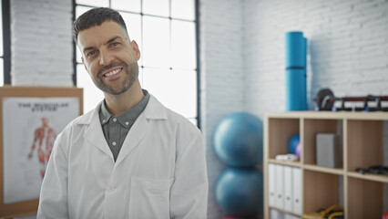 Confident hispanic male physiotherapist smiling in a bright rehab clinic interior.