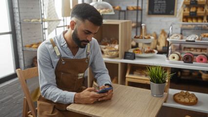 Handsome hispanic man with a beard and apron checking his phone in a cozy bakery shop filled with various baked goods