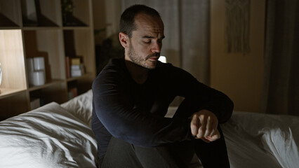 Fototapeta premium Pensive bald hispanic man with beard sitting in dark bedroom at night