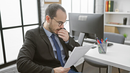 Fototapeta premium A focused man in glasses analyzes documents in a modern office with a computer, evoking professionalism and concentration.