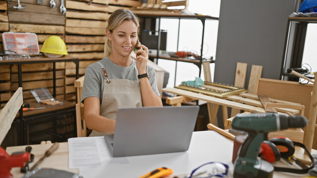 Young caucasian woman talks on phone at a carpentry workshop with laptop and tools.
