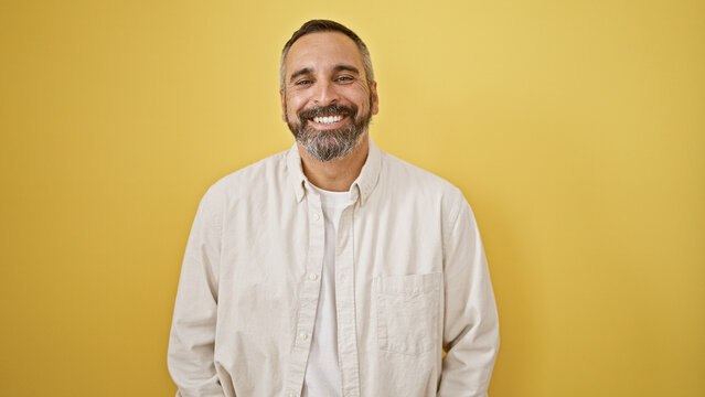 A smiling hispanic senior man with a beard and grey hair stands against a yellow isolated background wearing a white shirt.