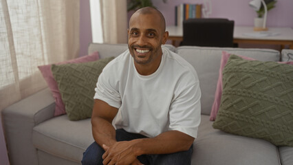 Young man sitting on a couch in a cozy living room, smiling warmly in a modern apartment interior with green pillows and soft lighting