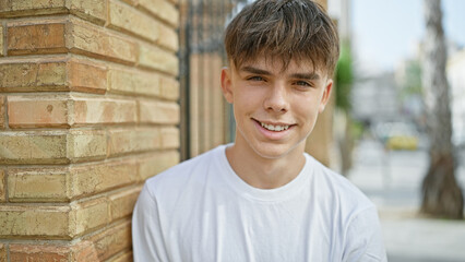 Obraz premium A young caucasian teen boy with blond hair, wearing a white shirt, smiles on a sunny urban street with a brick wall background.