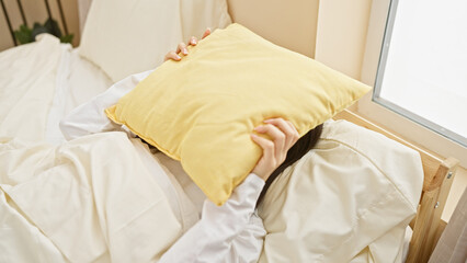 Young asian woman hiding face with pillow in bright bedroom setting