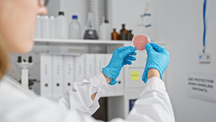 A young caucasian woman scientist examines a petri dish in a laboratory setting, representing healthcare research and clinical work.