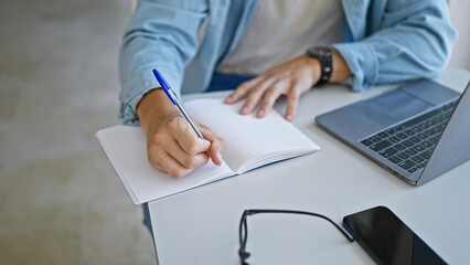 A young hispanic man studying in a university library, with a notebook, pen, laptop, and smartphone on the table.