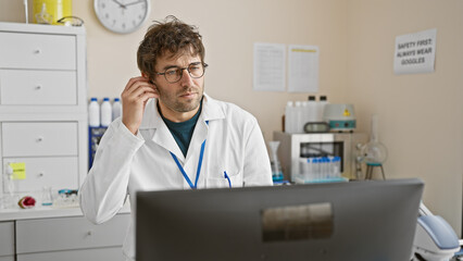 Young hispanic man with beard wearing wireless earphones and lab coat in clinic office, working at computer