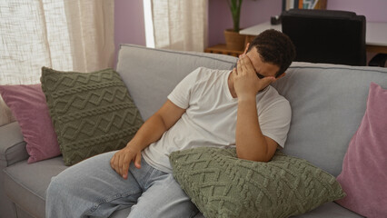 Young hispanic man sitting on a couch at home with his face buried in his hand, appearing stressed...