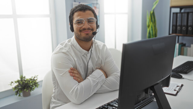Young man with crossed arms and headset smiling in a modern office, surrounded by computers and office supplies.