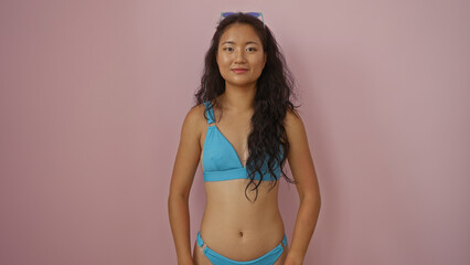 Young asian woman in a blue bikini stands isolated against a pink wall, exuding confidence and beauty.