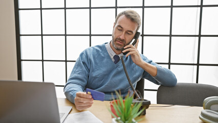 A professional middle-aged man engages in a phone conversation while holding a credit card in a...