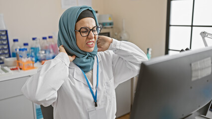 Fototapeta premium A middle-aged woman wearing a hijab experiences neck pain in a laboratory setting, embodying healthcare professionalism.