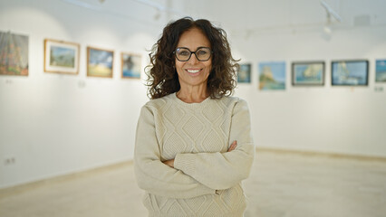 Confident middle-aged hispanic woman with glasses standing with arms crossed in an art gallery,...