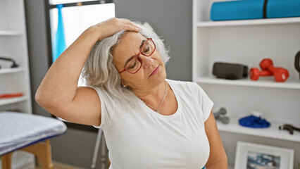 A mature woman with grey hair looks tired in a physical therapy clinic, indicating distress or exhaustion.