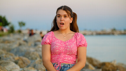 A somewhat chubby teenage girl sitting on the rocks by the seaside on a sunny summer day, showing surprised facial expressions