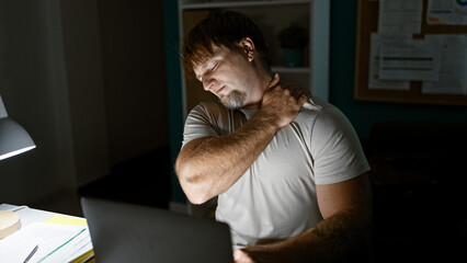 A young, bearded, blond man grimaces in pain as he holds his neck, working at a laptop in a dimly lit office room.