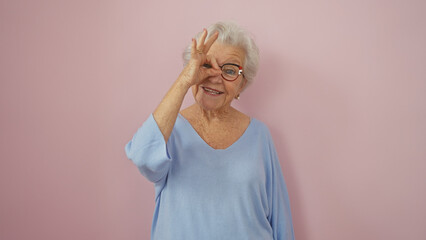 Smiling senior woman in blue sweater making okay gesture against a pink background.