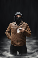 Drug dealer, dangerous hooded criminal man in black mask and brown leather jacket holding transparent plastic bag with drugs and dollar bills, on a dark background with smoke