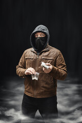 Drug dealer, dangerous hooded criminal man in black mask and brown leather jacket holding...