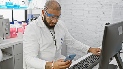 African american male scientist in a lab coat using a smartphone and a computer in a laboratory setting