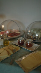 Breakfast buffet featuring various cheeses, jams, meats, olive oil, and condiments displayed under protective covers on a table indoors