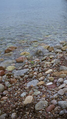 Rocky beach shoreline with pebbles and stones leading into translucent blue water under an overcast sky.