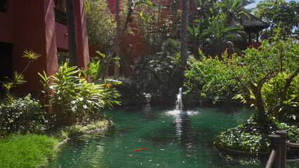 Tranquil pond with fountain centered in a lush tropical garden with green foliage and red buildings.