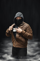 Drug dealer, dangerous hooded criminal man in black mask and brown leather jacket holding...