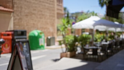 Outdoor cafe scene with blurred foreground, tables, and chairs, defocused view of bright sunny street with recycling bins and palm trees