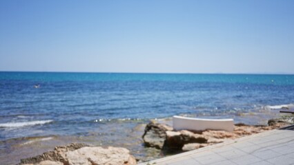 Blurred view of a seaside with rocks and clear blue water under a sunny sky in an out-of-focus, bokeh style.